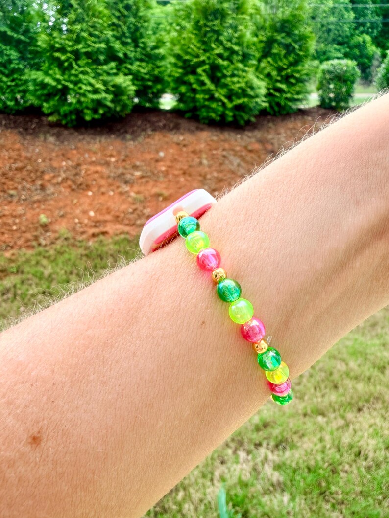 May include: A colorful beaded bracelet with pink, green, and yellow beads. The bracelet is accented with gold-colored spacers and a white and pink band. The bracelet is worn on a person's arm.