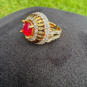 May include: A gold-tone ring featuring a central oval-shaped red stone surrounded by a halo of small clear stones. The band and setting are embellished with additional clear stones and a textured gold-tone finish, creating a layered appearance.