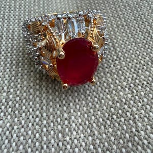 Puede incluir: Un anillo de tono dorado con una gran piedra central de color rojo intenso en forma ovalada sujeta por cuatro garras. La banda está decorada con motivos de hojas de metal dorado texturizado y acentuada con numerosas piedras blancas claras.