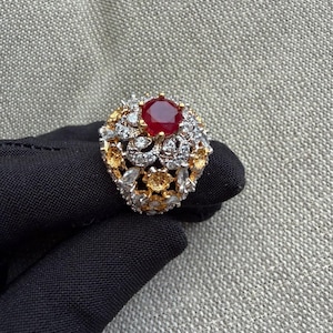 Puede incluir: Un anillo con una gran piedra central redonda de color rojo intenso, rodeada por un grupo de piedras blancas transparentes y piedras de tonos amarillos más pequeñas, engastadas en una montura metálica con un diseño calado de flores.