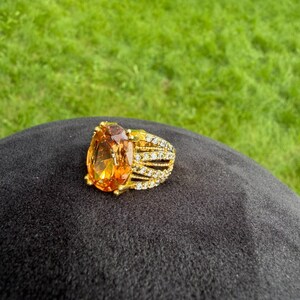 May include: A gold-toned ring featuring a large, oval-cut amber-coloured gemstone as the centrepiece. The band is split into multiple rows, each encrusted with small, clear, sparkling stones that extend along the sides of the central setting.
