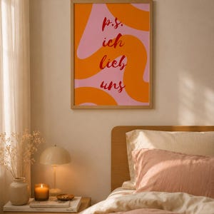 Könnte beinhalten: Ein rechteckiges Wandbild in einem hellen Holzrahmen mit einem rosafarbenen Hintergrund, wellenförmigen orangefarbenen Streifen und dem roten Text p.s. ich lieb uns in einer Schreibschrift. Das Bild hängt an einer Wand über einem Bett.