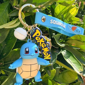 Op de afbeelding: Een blauwe Squirtle sleutelhanger met een goudkleurige ring, een witte balbedel en een blauwe polsband met de tekst "Squirtle". De sleutelhanger heeft ook een "Pokemon" logo bedel. De Squirtle figuur is blauw en bruin.