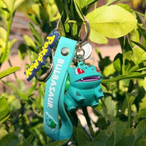 May include: A teal Bulbasaur keyring with a matching strap that reads "Bulbasaur" and "Pokemon". The Bulbasaur figure is green with red eyes and a smiling expression. The keyring is attached to a silver ring.