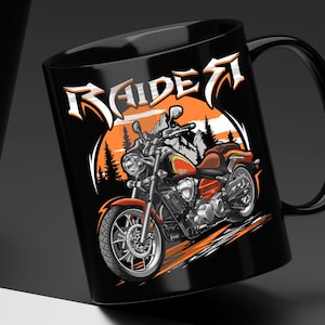 May include: A black ceramic mug featuring a graphic illustration of a motorcycle in orange, yellow, and silver tones. The design includes a mountain and forest background with the word "RAIDER" printed in a stylized white font above the motorcycle.