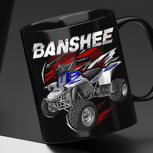 May include: A black ceramic mug featuring a graphic illustration of a blue and white all-terrain vehicle. The word BANSHEE is printed in large white capital letters above the vehicle, set against a background of abstract red and black shapes.