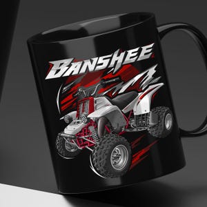 May include: A black ceramic mug featuring a graphic illustration of a white and red all-terrain vehicle. The word BANSHEE is printed in large, stylized white letters above the vehicle, set against a background of abstract red and white streaks.