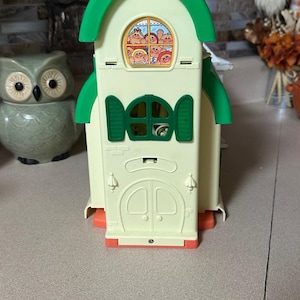 May include: A cream-colored plastic toy house featuring a bright green arched roof, green shutters, and a double door entrance. The upper window displays a colorful graphic of multiple cartoon characters with various hair colors and expressions.