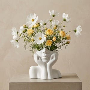 Peut inclure: Un vase en céramique blanche en forme de tête humaine reposant sur des mains, rempli d'un bouquet de fleurs cosmos blanches et de petites roses jaunes. Le vase présente une finition mate lisse et représente un visage aux yeux fermés.