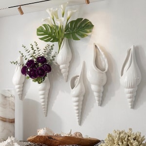 Peut inclure: Plusieurs vases muraux blancs en forme de coquillages en céramique, chacun avec un design unique. Certains vases contiennent des fleurs et de la verdure, notamment des roses pourpres, des callas et de grandes feuilles vertes. Les vases sont fixés sur un mur blanc, créant un thème côtier.