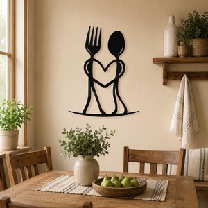 Op de afbeelding: Een zwart metalen wanddecoratie met het gestileerde silhouet van een vork en een lepel. De stelen van het bestek buigen naar binnen om een hartvorm in het midden te creëren, terwijl de onderkanten naar buiten uitlopen als een basis.