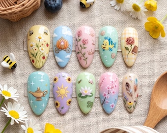 May include: A collection of ten almond-shaped nail art designs. Each nail features a unique, colorful design, including flowers, a pumpkin carriage, a genie lamp, and a sun. The nails are arranged on a neutral surface with decorative elements.
