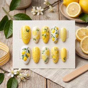 Pode incluir: Um conjunto de dez unhas artificiais com tema de limão. As unhas são pintadas com vários desenhos, incluindo limões inteiros, fatias de limão e ramos de limão, com fundos amarelos e brancos. As unhas estão dispostas em um cartão branco.