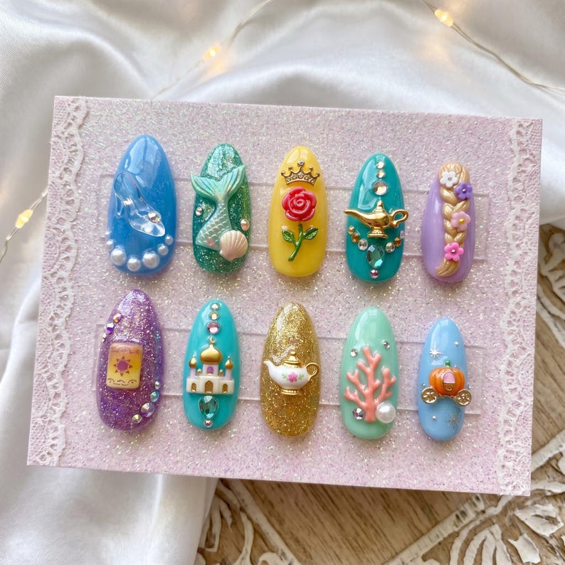May include: A set of ten artificial nails with various designs. The nails feature a range of colors, including blue, green, yellow, purple, gold, and teal. Each nail is decorated with miniature 3D embellishments, such as a mermaid tail, a rose, a lamp, and a castle.