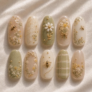 May include: A set of ten almond-shaped artificial nails with floral and bee designs. The nails feature a variety of colors, including beige, green, and gold, with intricate hand-painted details. The designs include daisies, honeycombs, and plaid patterns.