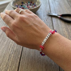 May include: A beaded bracelet featuring white square beads with black letters spelling CHICAGO, interspersed with red star-shaped beads, small silver-toned star charms, and smaller blue and red round beads on a clear elastic cord.
