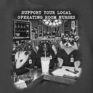 May include: A dark grey t-shirt featuring a graphic of an opossum, a skunk, and a raccoon dressed as medical staff in an operating room. The text reads SUPPORT YOUR LOCAL OPERATING ROOM NURSES. The animals wear scrubs and caps with medical symbols.