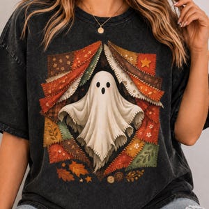 Puede incluir: Una camiseta gris carbón con lavado mineral que presenta un gráfico central de un fantasma blanco emergiendo de un diseño de patchwork. El diseño está compuesto por varios cuadrados de tela en tonos otoñales con motivos de hojas, estrellas y flores.