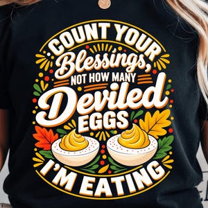 May include: A black t-shirt featuring a graphic design with the text "COUNT YOUR Blessings, NOT HOW MANY Deviled EGGS I'M EATING". The illustration includes two halves of a deviled egg with yellow yolks, surrounded by colorful autumn leaves in orange, red, and green.