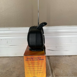 May include: A black portable AM/FM radio with a tall, extended telescopic metal antenna and a fold-down carrying handle. The radio sits atop its original orange and yellow cardboard box, which lists features including dynamic dual silver cone speakers.
