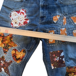 Peut inclure: Jean en denim bleu orné de divers empiècements en tissu en forme d'étoile, de cœur et de rectangle avec des motifs floraux, de renards et de champignons, fixés par des coutures blanches visibles, mesuré par une règle en bois de 60 cm.