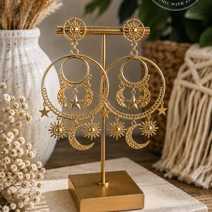 May include: A pair of gold-toned dangle earrings featuring celestial motifs, including suns, moons, and stars. The earrings are suspended from a gold-toned T-bar display stand. Each earring consists of a large hoop with smaller charms attached.