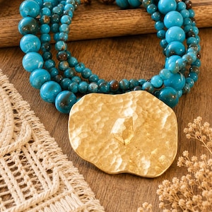 Puede incluir: Un collar de múltiples hilos con cuentas de color turquesa vibrante de varios tamaños y vetas de color marrón oscuro. El collar presenta un gran colgante de metal dorado martillado de forma irregular con un detalle central en relieve.