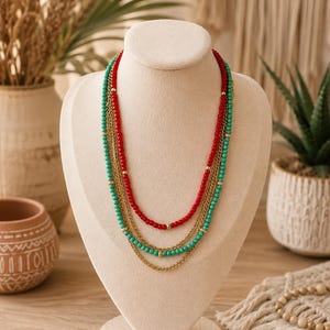 Puede incluir: Un collar de varias capas con una hilera de cuentas rojas, una hilera de cuentas turquesas y dos cadenas de metal dorado. El collar se exhibe en un busto de maniquí beige e incluye pequeñas cuentas espaciadoras de color dorado.