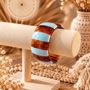 Peut inclure: Un large bracelet jonc orné de rayures verticales alternées bleu clair et en résine brune translucide à motif écaille de tortue. L'accessoire circulaire est présenté sur un support cylindrique beige. Un logo indique BOHO BADDIE CO.