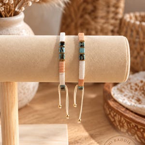 Puede incluir: Dos pulseras ajustables con una secuencia de cuentas planas en forma de disco en blanco, melocotón y turquesa, acentuadas con pequeños espaciadores metálicos. Cada pulsera incluye un cordón claro con dos pequeñas cuentas doradas.