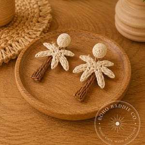 May include: A pair of palm tree earrings crafted from woven natural fibers. The earrings feature a round stud top, cream-colored palm fronds, and a brown trunk. A circular logo in the corner reads BOHO BADDIE CO. BOHO CHIC WITH ATTITUDE.