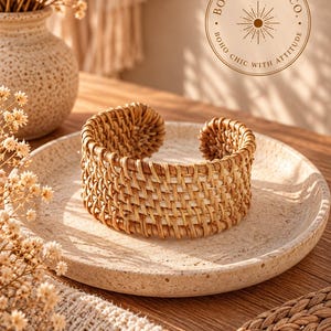 May include: A wide cuff bracelet crafted from woven light brown and tan rattan fibres. The bracelet features an open-ended design with a textured, basket-weave pattern. The circular logo BOHO BADDIE CO. BOHO CHIC WITH ATTITUDE is visible above.