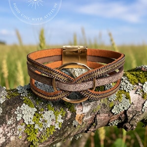 May include: A multi-strand bracelet featuring brown leather-like bands, metallic gold-tone chains, and a central gold-tone circular ring. The bracelet is secured with a polished gold-tone magnetic clasp. The text BOHO BADDIE CO. BOHO CHIC WITH ATTITUDE is visible.