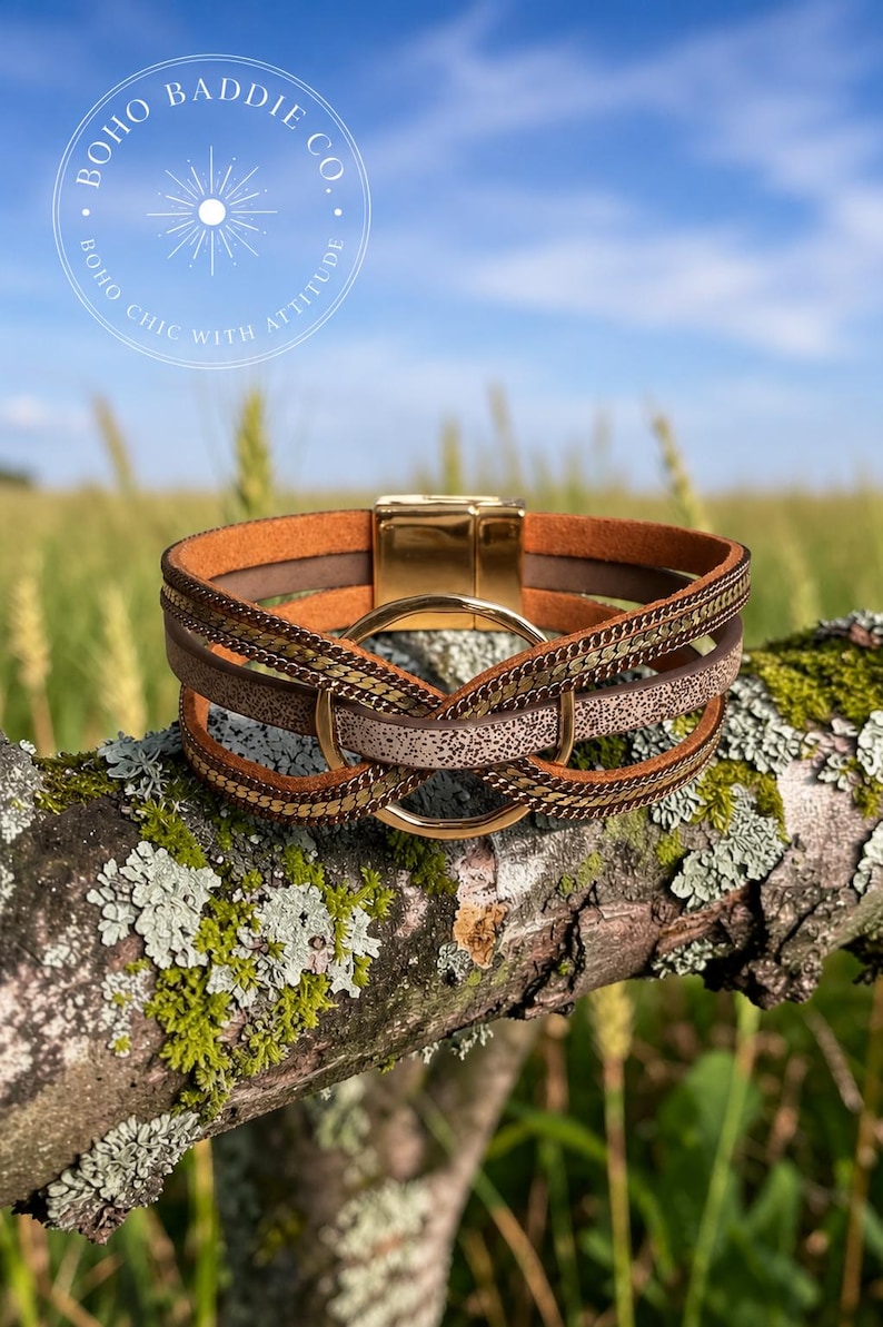 May include: A multi-strand bracelet featuring brown leather-like bands, metallic gold-tone chains, and a central gold-tone circular ring. The bracelet is secured with a polished gold-tone magnetic clasp. The text BOHO BADDIE CO. BOHO CHIC WITH ATTITUDE is visible.