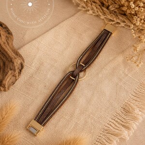 May include: A multi-strand bracelet featuring brown faux leather strips and metallic gold-tone chains. The strands are looped through a central gold-tone metal ring and secured with a rectangular gold-tone magnetic clasp at each end.