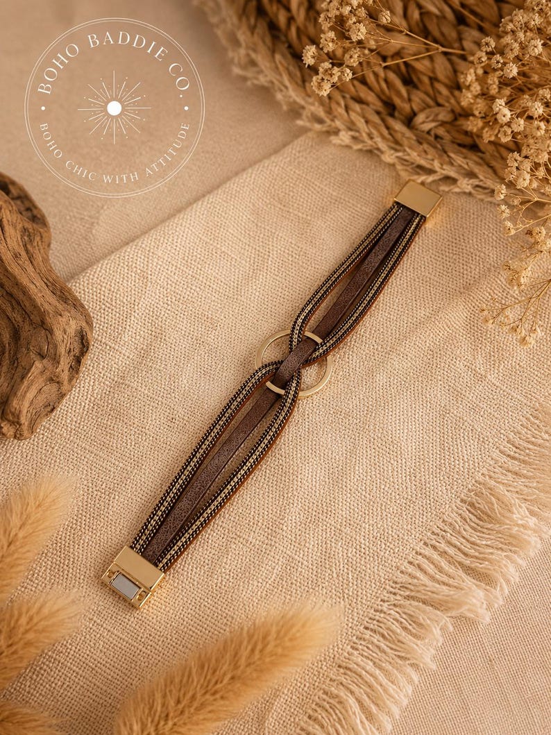 May include: A multi-strand bracelet featuring brown faux leather strips and metallic gold-tone chains. The strands are looped through a central gold-tone metal ring and secured with a rectangular gold-tone magnetic clasp at each end.