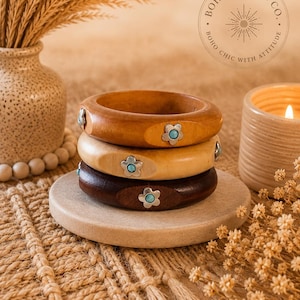 May include: A stack of three wooden bangles in varying shades of brown, each featuring a silver-toned flower-shaped inlay with a small, round, light blue stone at the centre. The bracelets are arranged vertically on a circular stone base.