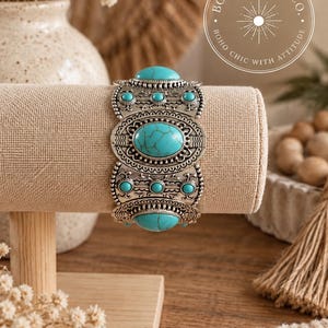 May include: A silver-toned cuff bracelet featuring multiple oval and circular turquoise-coloured stones with dark veining. The metal surface displays intricate embossed patterns and beaded edges. A circular logo reads BOHO BADDIE CO. BOHO CHIC WITH ATTITUDE.