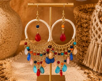 May include: A pair of gold-tone hoop earrings featuring white cord wrapping, accented with dangling multi-colored beads in shades of red, blue, orange, and translucent pink. The earrings hang from a gold-tone metal T-shaped display stand.