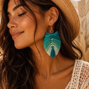 Puede incluir: Un par de pendientes en forma de hoja hechos de hilo de flecos en degradado azul verdoso, turquesa y blanco. Los pendientes cuentan con un gancho de metal dorado y un diseño central de cordón anudado que imita las venas de una hoja.