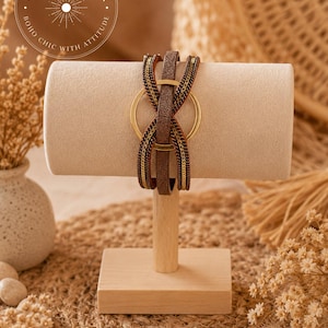 May include: A multi-strand wrap bracelet featuring a central gold-toned metal ring, a brown textured leather-like strap, and two flanking metallic chain strands in a dark bronze finish, all secured together in a layered, crisscross design.