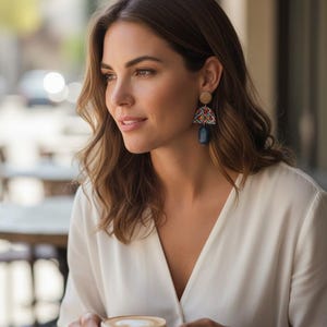 May include: A woman wearing decorative earrings with a wooden stud, a colourful patterned section, and a dark blue teardrop. She is wearing a white blouse and holding a cup of coffee with a white saucer.