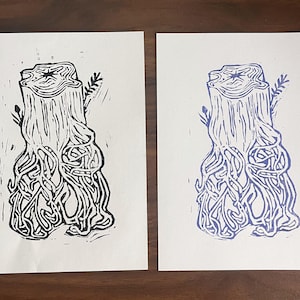 Op de afbeelding: Twee rechthoekige witte papieren afdrukken met een linosnede-illustratie van een boomstronk met blootliggende wortels. De linker afdruk gebruikt zwarte inkt, terwijl de rechter afdruk blauwe inkt gebruikt. Beide tonen dezelfde houtnerf.