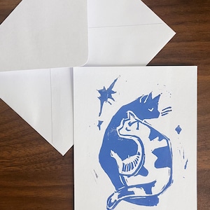 Op de afbeelding: Een witte wenskaart met een blauwe linosnede van twee katten die samen zitten, &eacute;&eacute;n effen blauw en &eacute;&eacute;n met blauwe vlekken, vergezeld door kleine stervormen, samen met een bijpassende witte envelop.