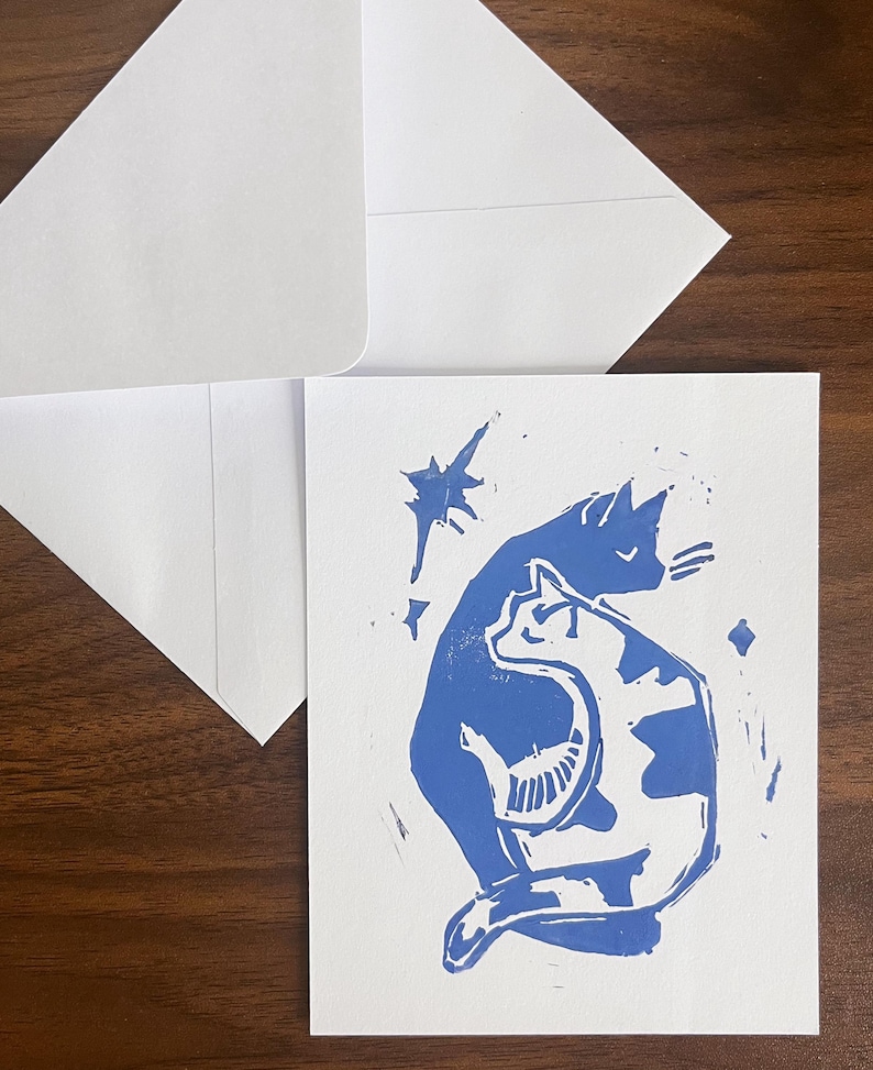 Op de afbeelding: Een witte wenskaart met een blauwe linosnede van twee katten die samen zitten, &eacute;&eacute;n effen blauw en &eacute;&eacute;n met blauwe vlekken, vergezeld door kleine stervormen, samen met een bijpassende witte envelop.