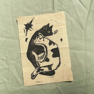 May include: A black and white linocut print of two cats embracing, with a star and diamond shapes, on a beige fabric. The print is on a piece of fabric with frayed edges, set against a green background.