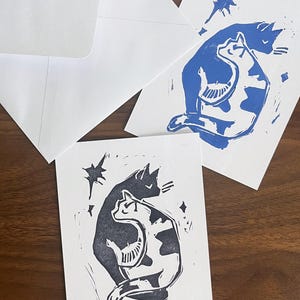 May include: Two rectangular greeting cards with white envelopes, each featuring a block print illustration of two cats sitting together. One card displays the cat design in blue ink, while the other card features the same design in dark gray ink.