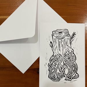 Op de afbeelding: Een witte wenskaart met een zwarte inktblokdruk van een boomstronk met blootliggende, ingewikkelde wortels die naar beneden uitsteken. De kaart wordt vergezeld door een bijpassende blanco witte envelop op een glad houten oppervlak.
