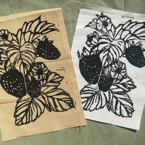 May include: Two fabric squares with a black and white strawberry plant design. One square is tan, the other is white. The design includes strawberries, leaves, and flowers. The fabric has a raw edge.