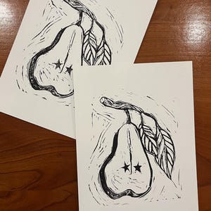 Op de afbeelding: Twee rechthoekige witte papieren prints met een zwarte linoleumsnede-illustratie van een gehalveerde peer. De peer heeft een steeltje, twee bladeren en twee kleine stervormen die in het midden van de vrucht zijn uitgesneden.